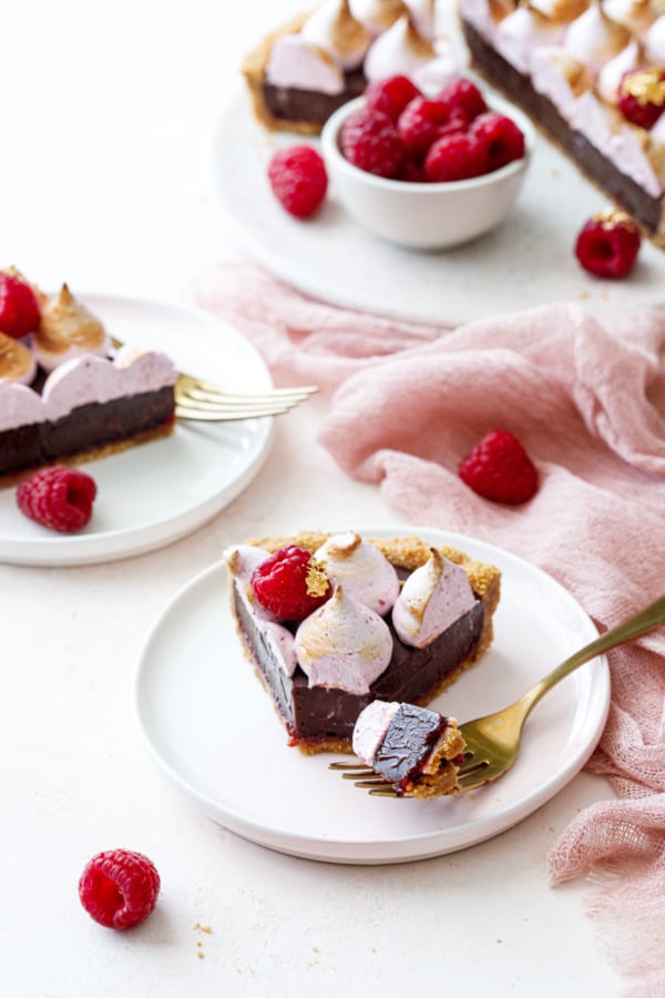 Slice of Chocolate Raspberry S'mores Tart with a bite on a fork, second plate and full tart on cake plate in the background.