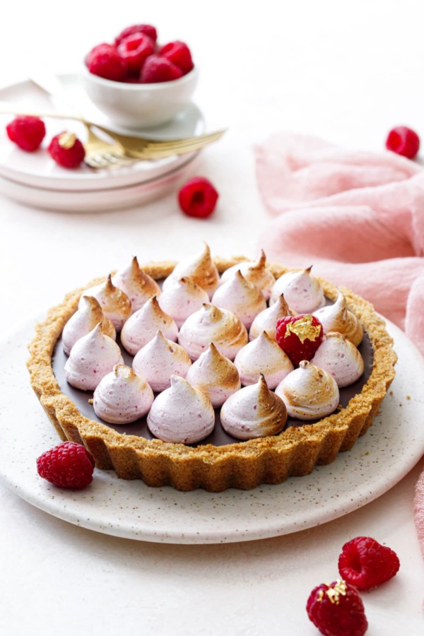 Chocolate Raspberry S'mores Tart on a light pink serving dish, pink linen and fresh raspberries in the background