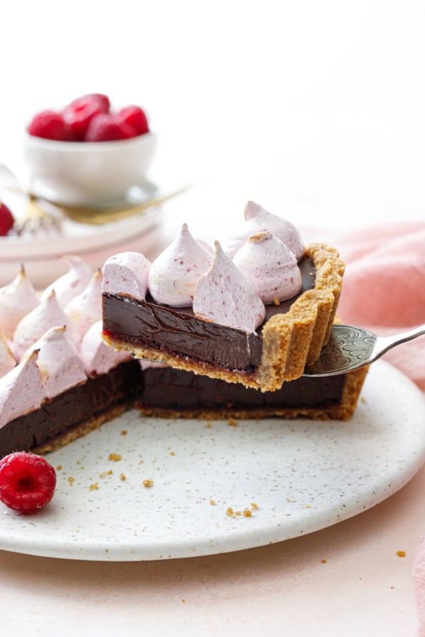 Silver serving utensil lifting a perfectly cut wedge of Chocolate Raspberry S'mores Tart; pink cake plate and fresh raspberries in the background.