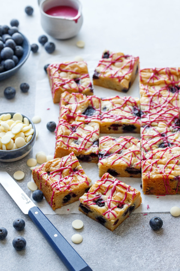 Squares of Blueberry White Chocolate Blondies drizzled with a hot pink glaze, on a gray background with bowls of white chips, blueberries, and a tiny pitcher of glaze in the background.