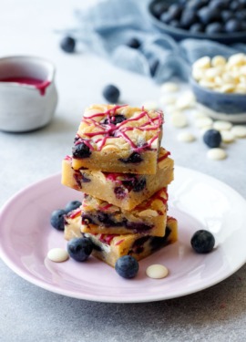 Stack of Blueberry White Chocolate Blondies cut into squares on a purple plate, gray background with raw ingredients and a pot of glaze in the background.