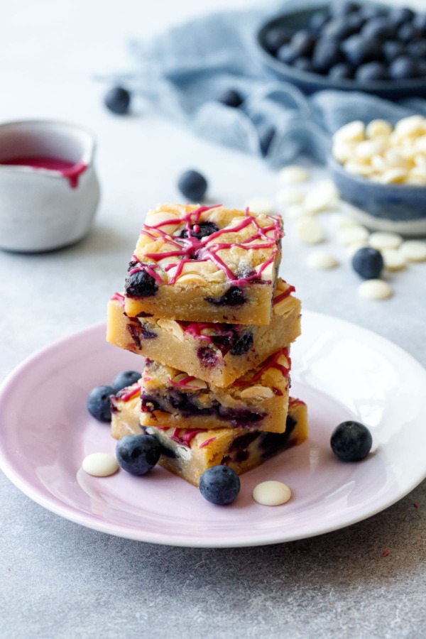Stack of Blueberry White Chocolate Blondies cut into squares on a purple plate, gray background with raw ingredients and a pot of glaze in the background.