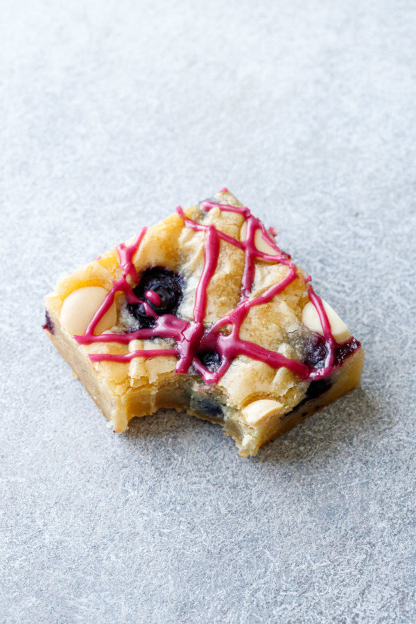One square of Blueberry White Chocolate Blondies on a gray background, with a perfect bite taken out of the corner.