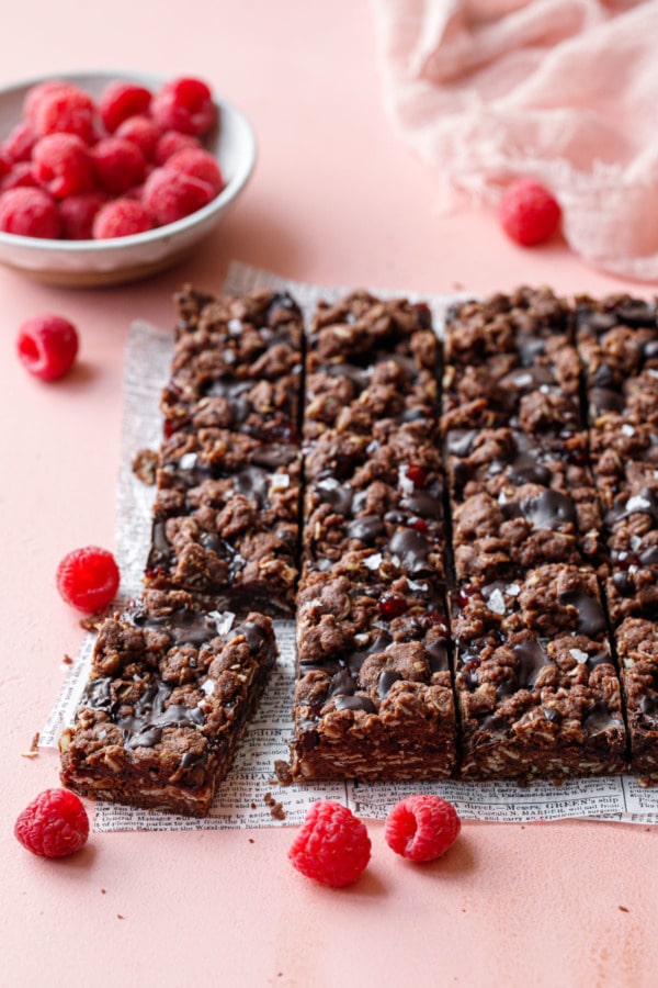 Rectangular cut Chocolate Raspberry Crumb Bars on parchment, pink background with fresh raspberries
