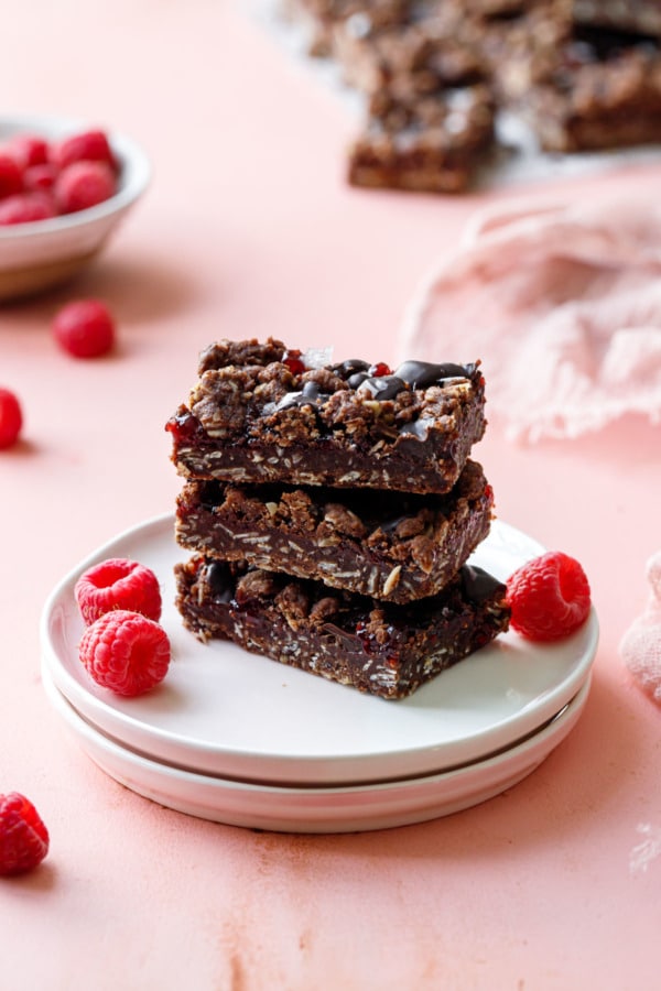 Small plate with stack of 3 rectangular-cut Chocolate Raspberry Crumb Bars, with pink background and fresh raspberries.