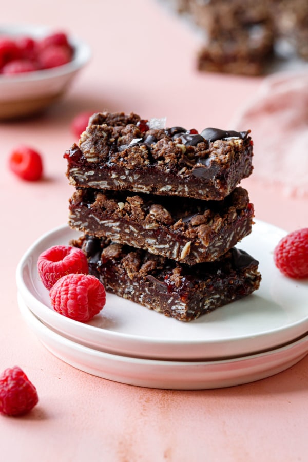 Stack of three Chocolate Raspberry Crumb Bars on small plates with fresh raspberries and bowl of raspberries in the background.