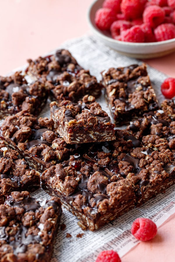 Chocolate Raspberry Crumb Bars on newsprint parchment paper, stacked haphazardly with a bowl of fresh raspberries.