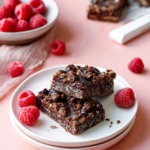 Two Chocolate Raspberry Crumb Bars on a plate with bowl of raspberries and more bars in the background.