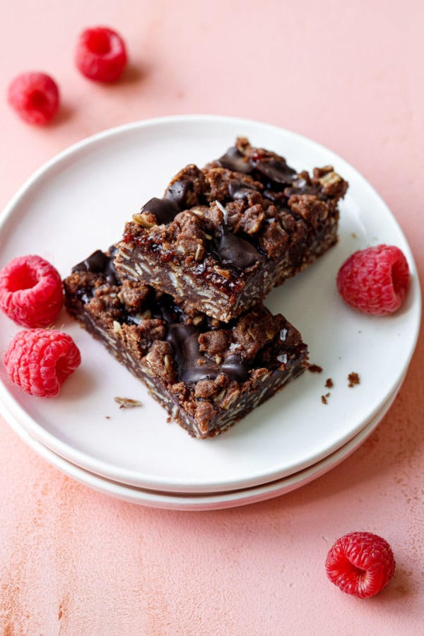 Two Chocolate Raspberry Crumb Bars arranged crosswise on a plate, with fresh raspberries on a pink background.