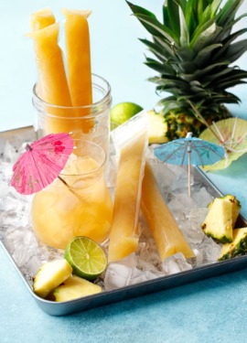 Aqua blue background with metal tray filled with ice, and mai tai cocktail, bright orange ice pops, and pineapple pieces