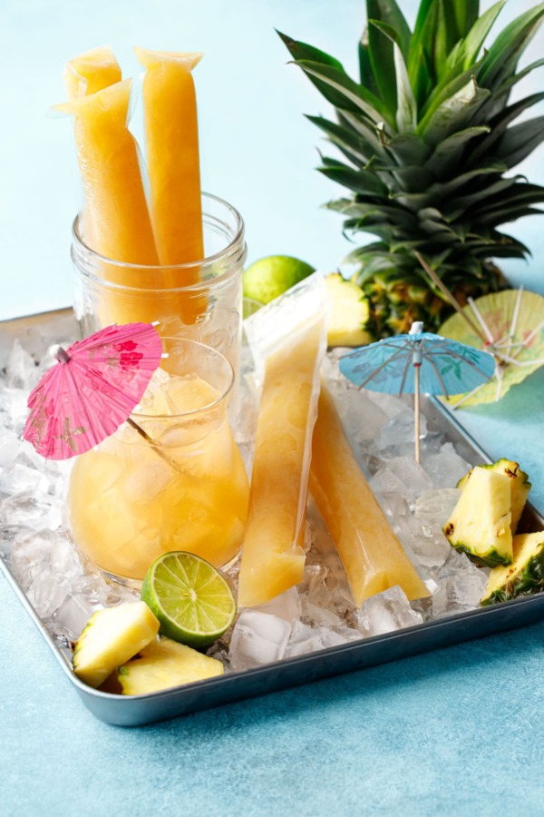 Aqua blue background with metal tray filled with ice, and mai tai cocktail, bright orange ice pops, and pineapple pieces