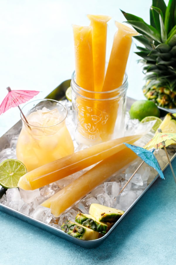 Frozen Mai Tai Slushy Pops on a metal tray with crushed ice and mai tai cocktail with pink umbrella.