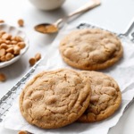 Three Salted Peanut Butter Chip Cookies on a wire rack with crinkled parchment, bowl of peanut butter and dish of peanut butter chips in the background.