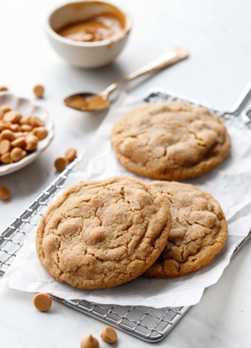 Three Salted Peanut Butter Chip Cookies on a wire rack with crinkled parchment, bowl of peanut butter and dish of peanut butter chips in the background.