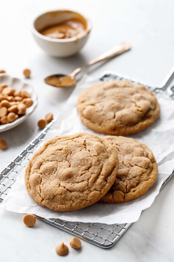 Three Salted Peanut Butter Chip Cookies on a wire rack with crinkled parchment, bowl of peanut butter and dish of peanut butter chips in the background.