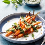 Blue and white ceramic platter with pile of Roasted Carrots, drizzled with Basil and Carrot Top Pesto on a blue background.