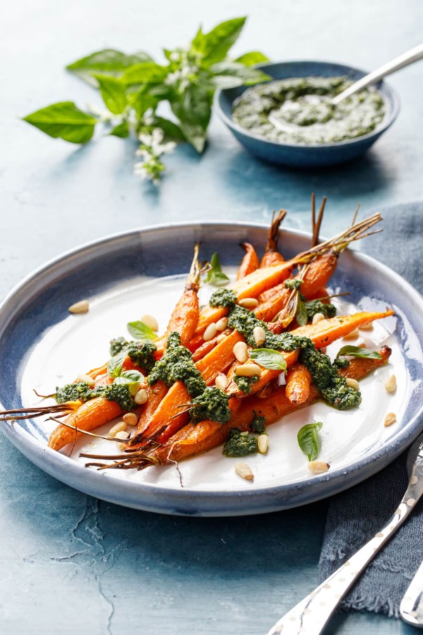 Blue and white ceramic platter with pile of Roasted Carrots, drizzled with Basil and Carrot Top Pesto on a blue background.