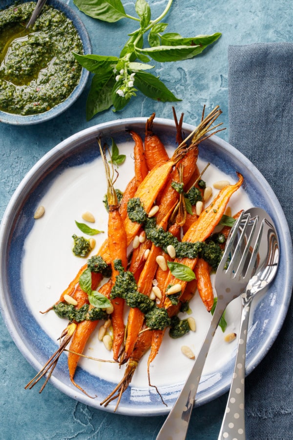 Plate with Roasted Carrots and silver serving utensils, bowl of Basil and Carrot Top Pesto on the side with sprig of fresh basil.