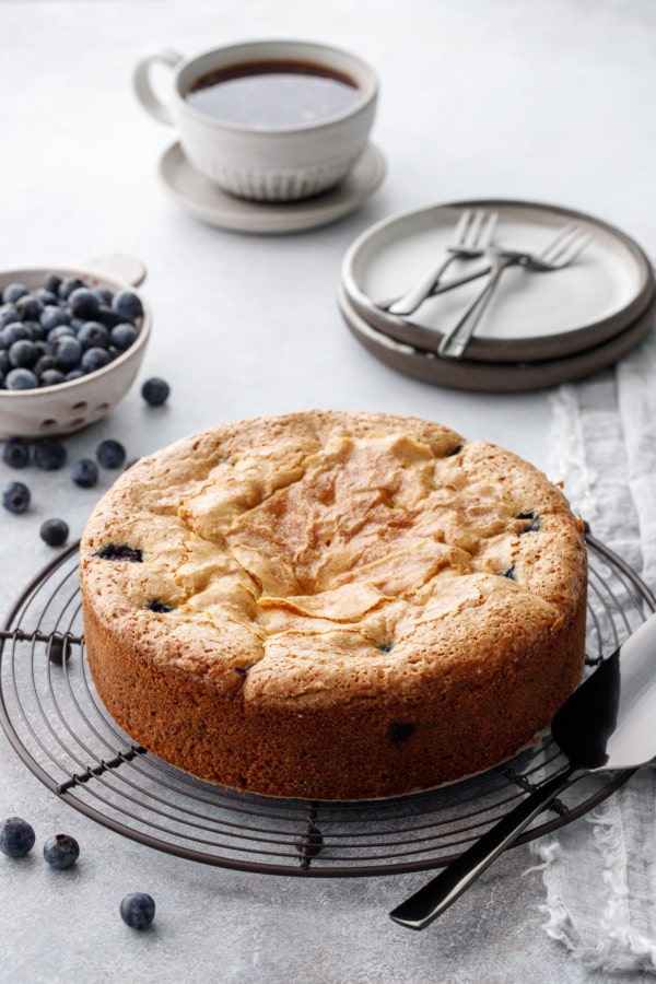 Blueberry Cream Cheese Coffee Cake on a wire rack, gray background with bowl of berries and cup of coffee in the background.