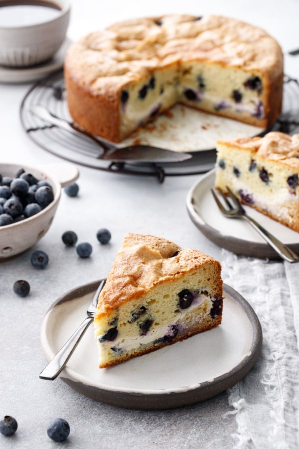 Slice of Blueberry Cream Cheese Coffee Cake on a ceramic plate, bowl of blueberries and cup of coffee in the background.