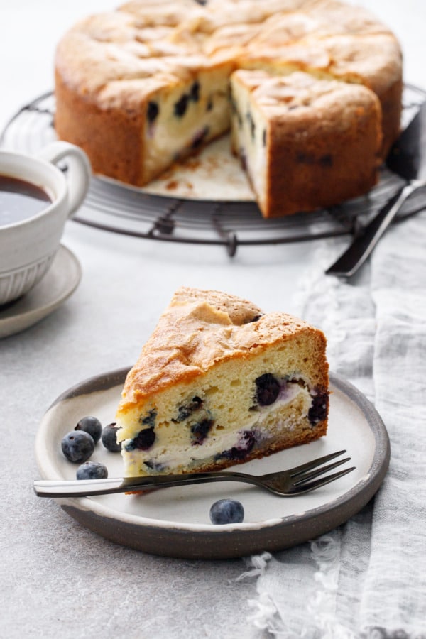 Slice of Blueberry Cream Cheese Coffee Cake on a ceramic plate with blueberries, a clear layer of cream cheese runs through the middle of the cake.