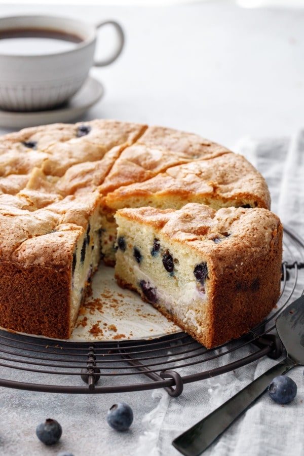 Sliced Blueberry Cream Cheese Coffee Cake on a wire rack, showing the visible cream cheese layer that runs through the middle of the cake; cup of coffee in the background.