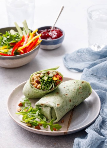 One cut Curried Chicken, Chickpea & Arugula Wrap on a plate, bowls of ingredients blurred in the background.