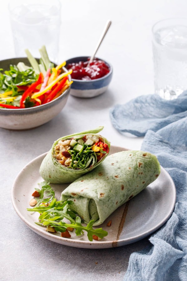 One cut Curried Chicken, Chickpea & Arugula Wrap on a plate, bowls of ingredients blurred in the background.