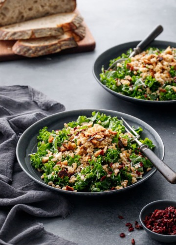 Two dark gray bowls of Kale & Farro Salad with Sourdough Breadcrumbs on a dark gray background, loaf of sourdough bread in the background.