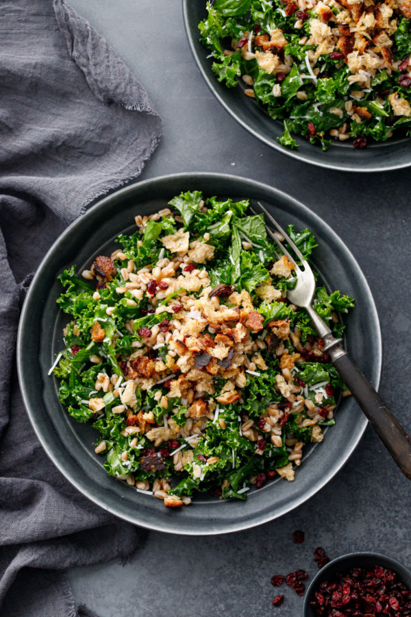 Overhead, dark gray bowls with Kale & Farro Salad with Sourdough Breadcrumbs and a vintage fork, on a charcoal gray background with linen.