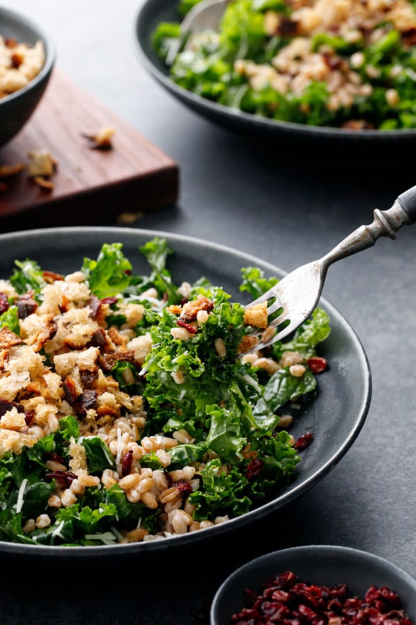 Fork lifting a bite of Kale & Farro Salad with Sourdough Breadcrumbs, with another bowl and bowl of breadcrumbs in the background.