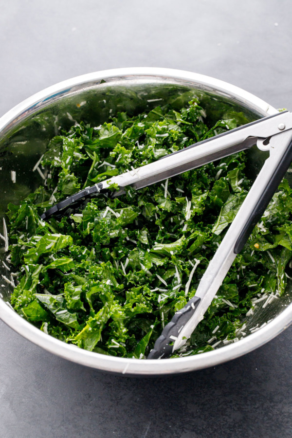 Metal mixing bowl, salad tongs tossing kale with dressing and pecorino cheese.
