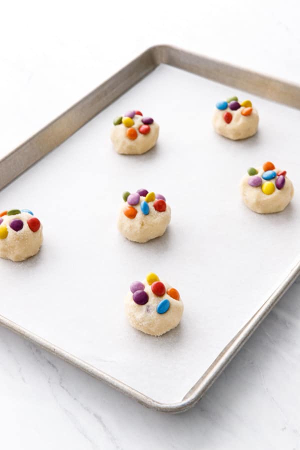 Cookie sheet lined with parchment, with six balls of sugar cookie dough topped with colorful M&Ms candies.