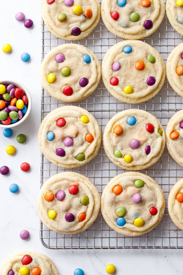 Overhead, wire rack with even rows of baked M&M Sugar Cookies, with a small bowl of M&Ms and more candies scattered around.