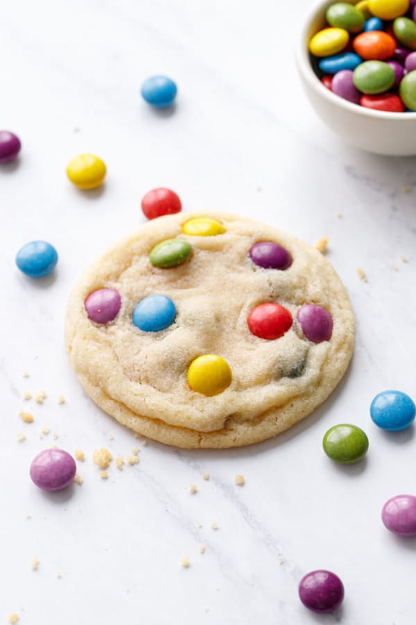 Focus on a single perfect M and M Sugar Cookie, with scattered M&Ms around on a white marble background.