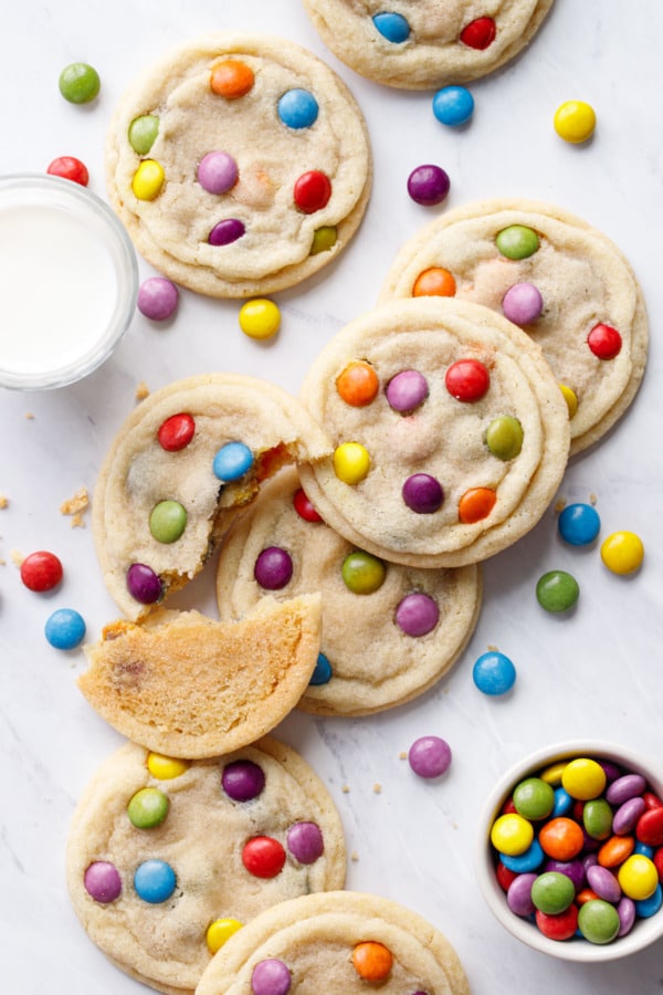 Overhead, messy scattered M&M Sugar Cookies on marble background, one cookie broken in half, glass of milk and scattered M&Ms candies.