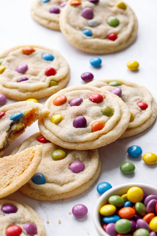 Messy arrangement of M&M Sugar Cookies on marble background, with bowl of candies and one cookie broken in half to show texture.