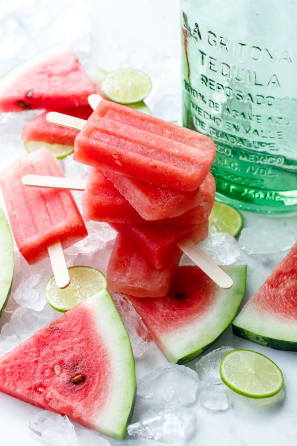 Stack of 4 Tequila Watermelon Popsicles, with crushed ice, sliced watermelon and lime, and bottle of tequila in the background.