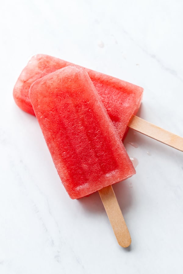 Two Tequila Watermelon Popsicles stacked on white marble background, close enough to show the crystalline frozen texture.