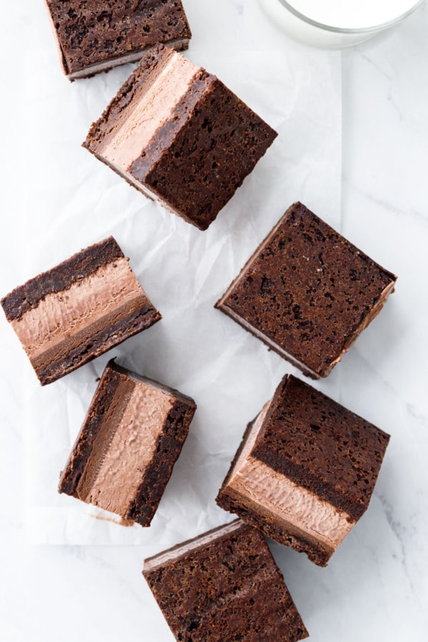 Overhead, random arrangement of Malted Fudge Brownie Ice Cream Sandwiches cut into squares, on a crinkled piece of parchment with glass of milk.