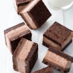 Random arrangement of Malted Fudge Brownie Ice Cream Sandwiches cut into squares, on a crinkled piece of parchment with glass of milk.