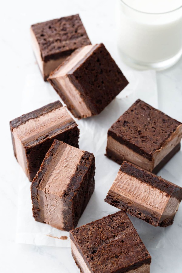 Random arrangement of Malted Fudge Brownie Ice Cream Sandwiches cut into squares, on a crinkled piece of parchment with glass of milk.