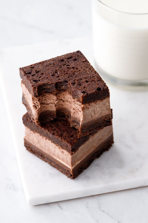 Stack of two Malted Fudge Brownie Ice Cream Sandwiches, one with a bite taken out of it, on a marble board with glass of milk in the background.