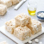 Stack of Olive Oil Rice Krispie Treats on a marble trivet, gray background with mini marshmallows and glass of olive oil in the background.