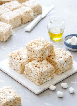 Stack of Olive Oil Rice Krispie Treats on a marble trivet, gray background with mini marshmallows and glass of olive oil in the background.