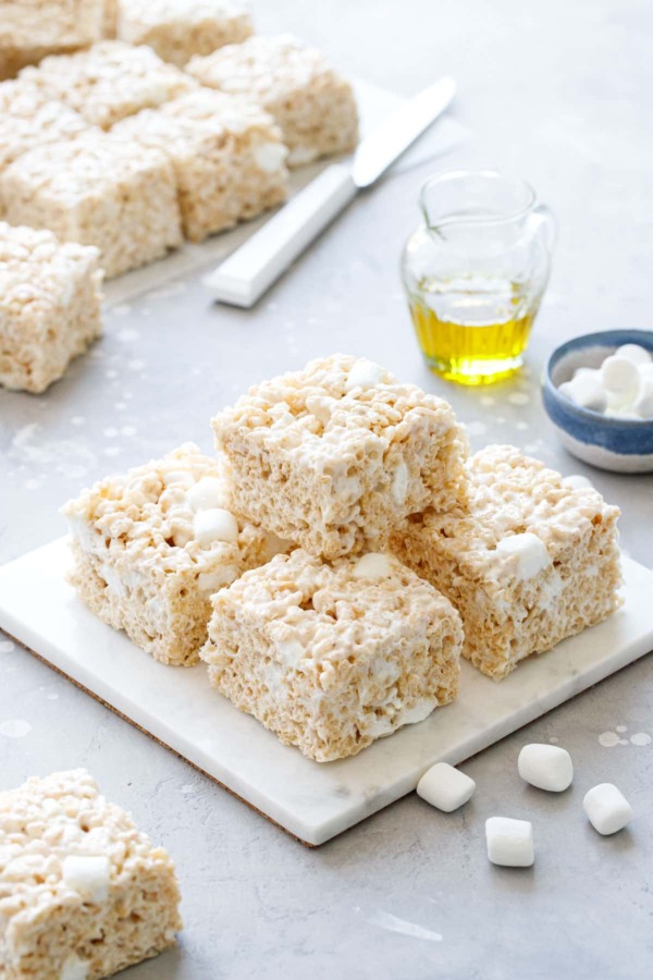 Stack of Olive Oil Rice Krispie Treats on a marble trivet, gray background with mini marshmallows and glass of olive oil in the background.