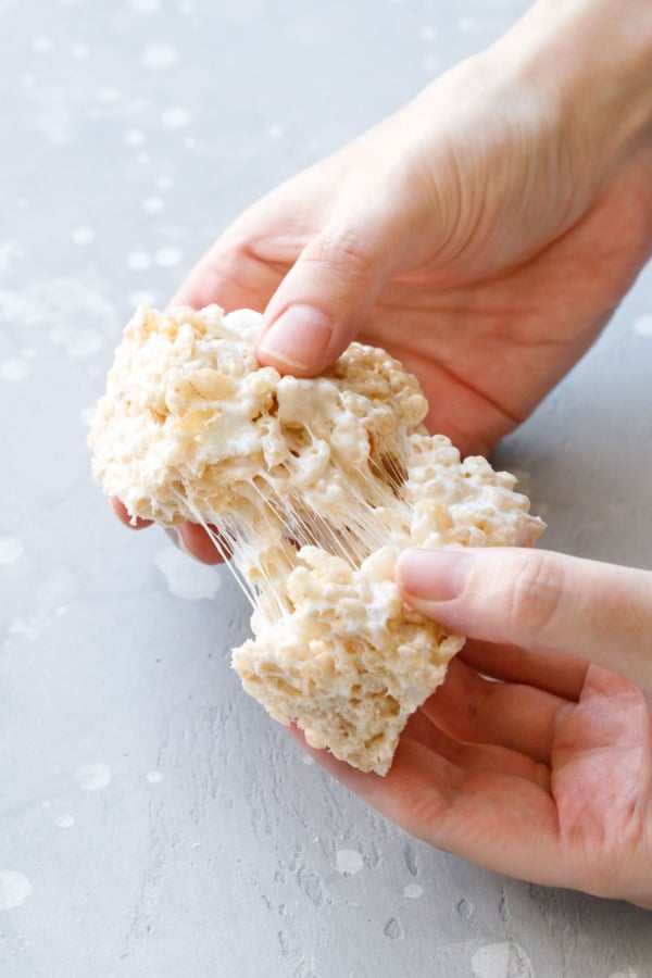 Hands pulling apart one square Olive Oil Rice Krispie Treat, showing threads of stretchy marshmallow.