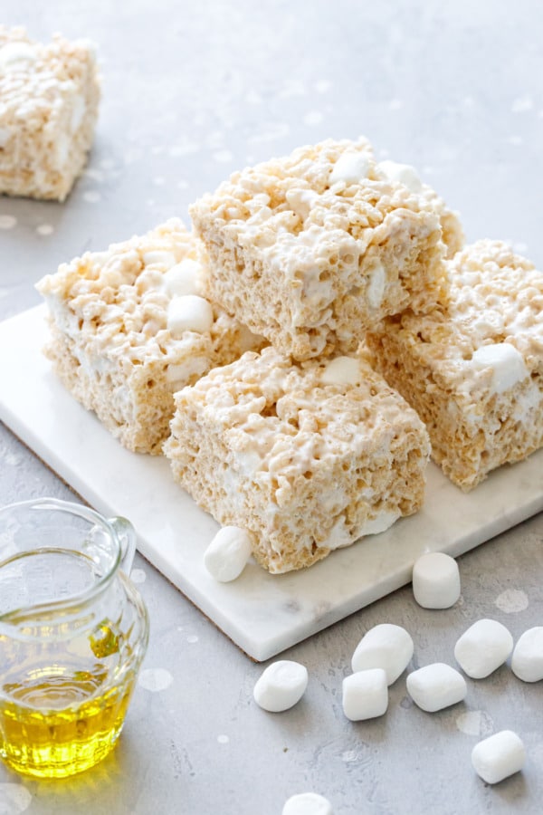 Stack of Olive Oil Rice Krispie Treats on a marble trivet, gray background with mini marshmallows and glass of olive oil in the background.