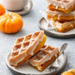 Two plates with stacks of Pumpkin Donut Waffles with a visibly drippy Vanilla Cardamom Glaze, mini pumpkins and cup of coffee in the background.