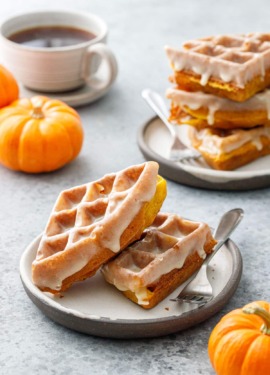 Two plates with stacks of Pumpkin Donut Waffles with a visibly drippy Vanilla Cardamom Glaze, mini pumpkins and cup of coffee in the background.